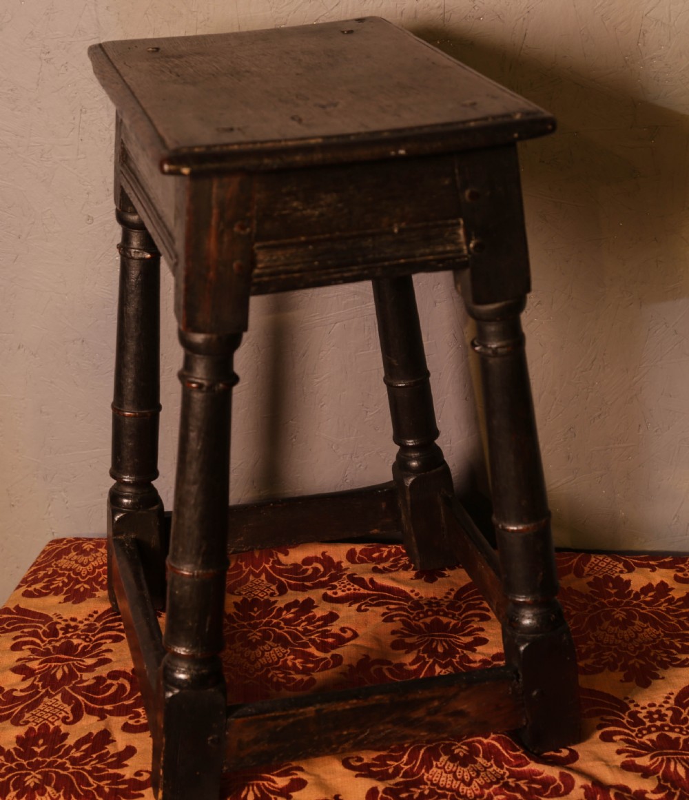 A seventeenth-century dark oak joint stool featuring turned legs, lower perimeter stretchers, and a flat top secured with wooden pegs. View V