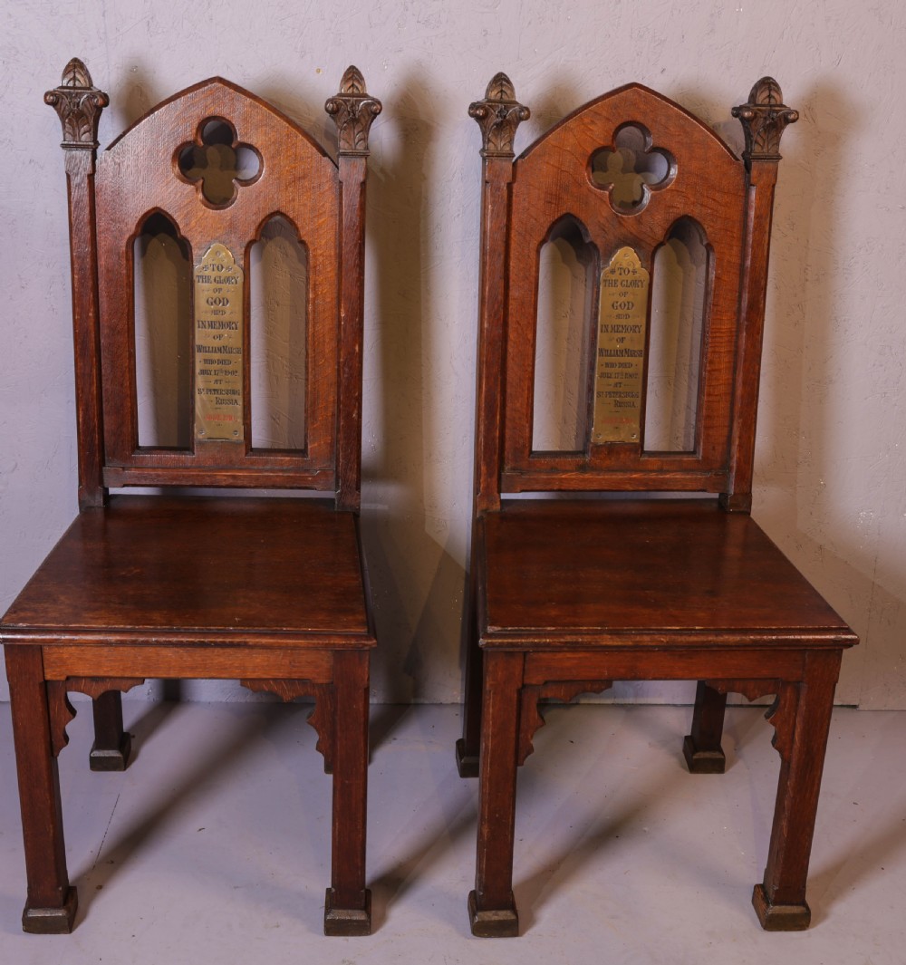 A pair of dark oak Gothic Revival church chairs featuring pointed arch backs, carved trefoil tracery, decorative corner finials, and central brass memorial plaques.