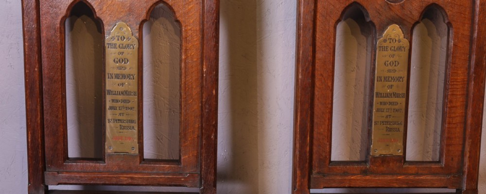 A pair of dark oak Gothic Revival church chairs featuring pointed arch backs, carved trefoil tracery, decorative corner finials, and central brass memorial plaques. View II
