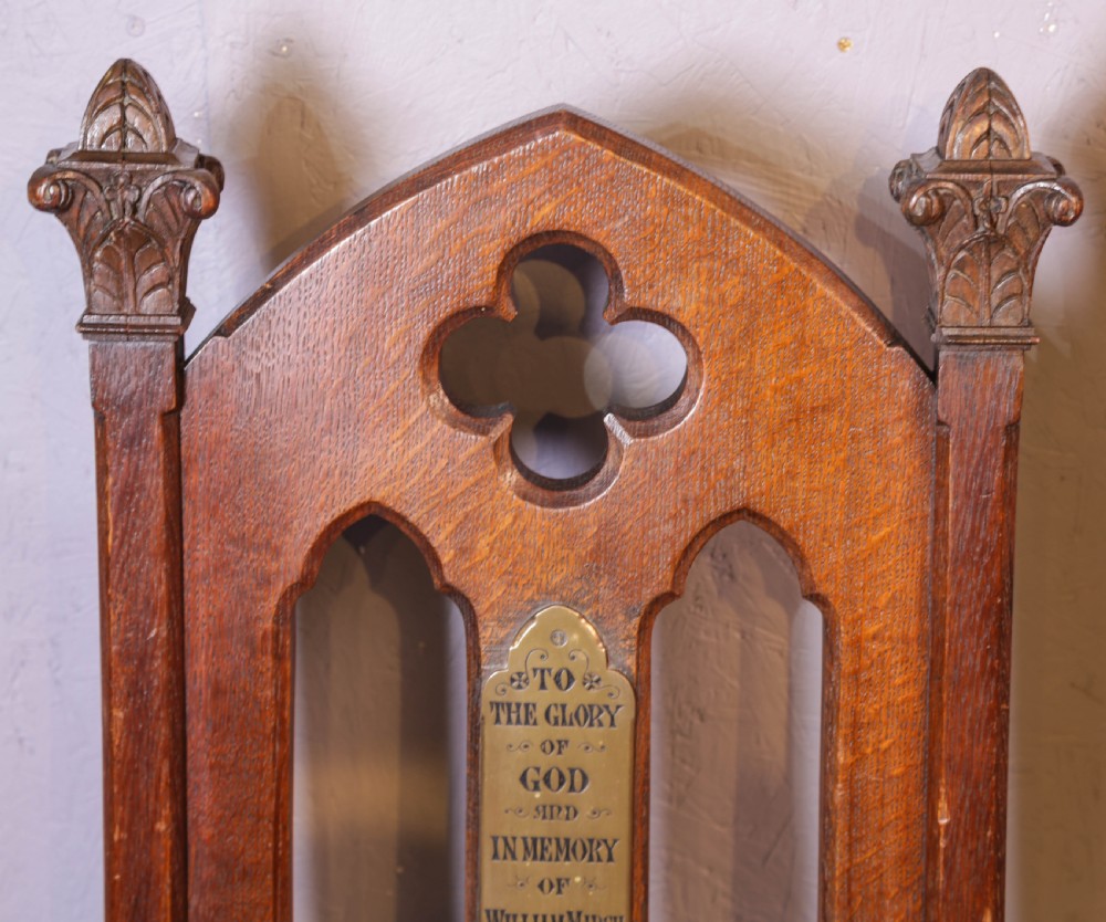 A pair of dark oak Gothic Revival church chairs featuring pointed arch backs, carved trefoil tracery, decorative corner finials, and central brass memorial plaques. View V