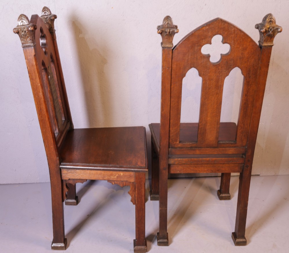 A pair of dark oak Gothic Revival church chairs featuring pointed arch backs, carved trefoil tracery, decorative corner finials, and central brass memorial plaques. View X
