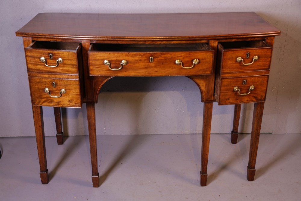 A Georgian mahogany bowfront sideboard featuring a central arched aperture, drawer storage with brass swan-neck handles, and square tapered legs. View IV
