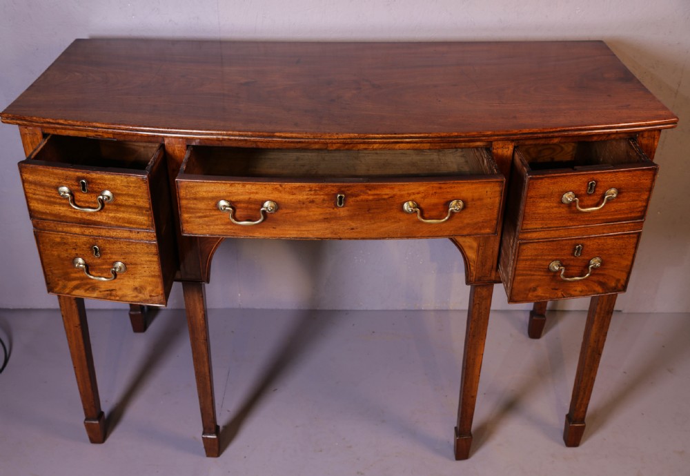 A Georgian mahogany bowfront sideboard featuring a central arched aperture, drawer storage with brass swan-neck handles, and square tapered legs. View VI