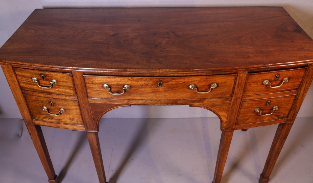 A Georgian mahogany bowfront sideboard featuring a central arched aperture, drawer storage with brass swan-neck handles, and square tapered legs. View VII
