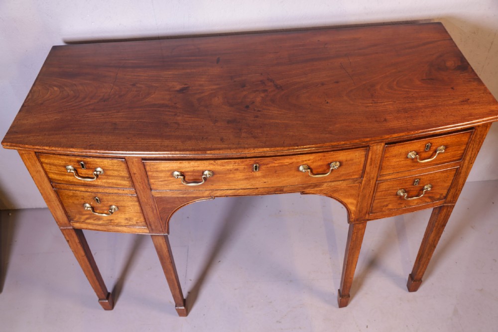 A Georgian mahogany bowfront sideboard featuring a central arched aperture, drawer storage with brass swan-neck handles, and square tapered legs. View VIII