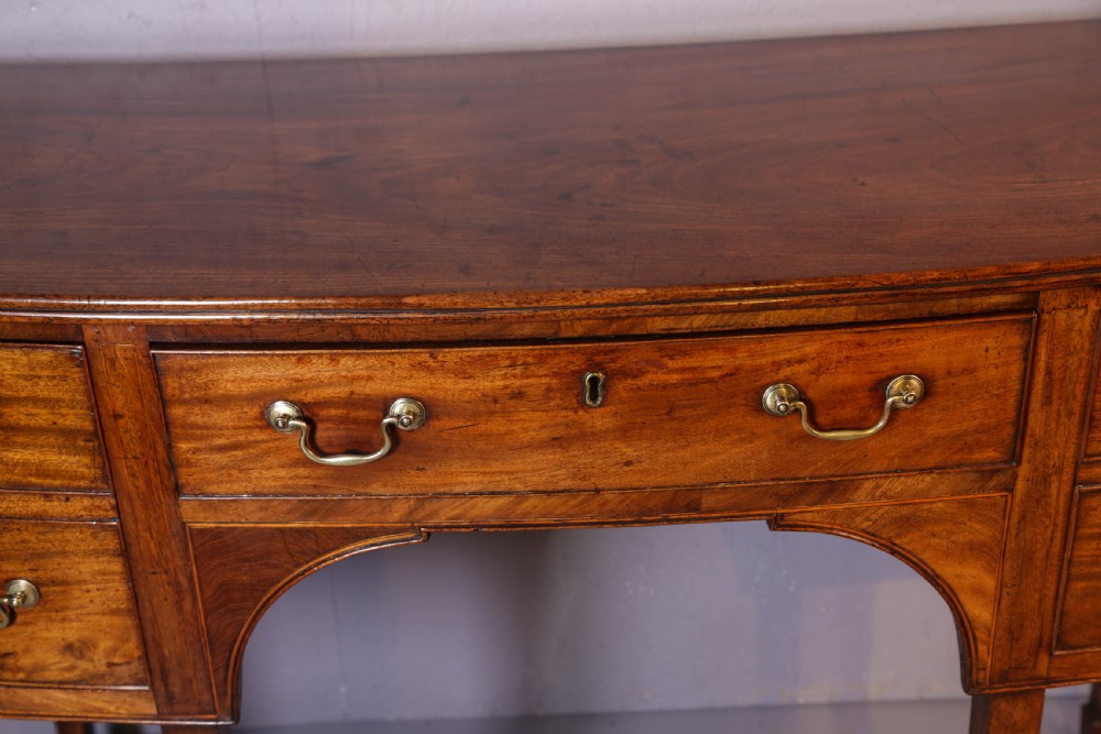 A Georgian mahogany bowfront sideboard featuring a central arched aperture, drawer storage with brass swan-neck handles, and square tapered legs. View XI