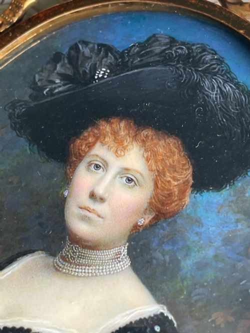 fig 1. An oval Edwardian portrait miniature depicting a woman with reddish hair in a black feather hat, encased in a gilt brass frame with a ribbon bow crest.