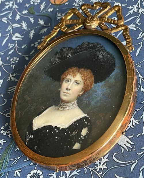 fig 5. An oval Edwardian portrait miniature depicting a woman with reddish hair in a black feather hat, encased in a gilt brass frame with a ribbon bow crest.
