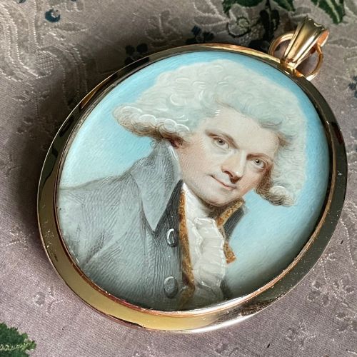 fig 1. An oval Georgian portrait miniature depicting a gentleman with a white powdered wig and grey coat, set within a gold frame with a top loop.