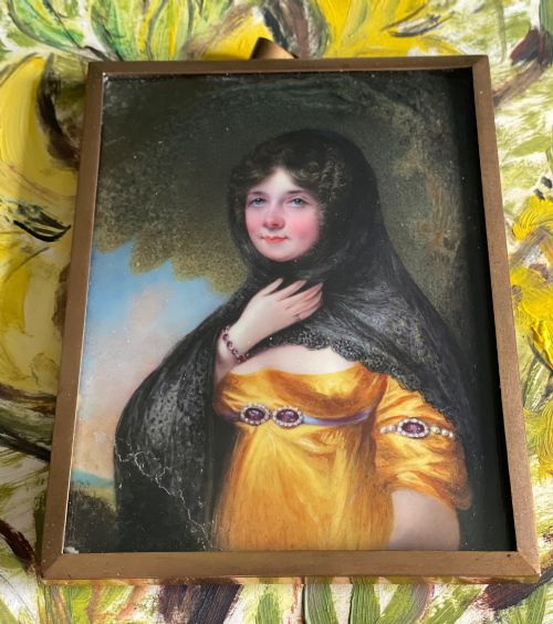 fig 1. A Georgian enamel portrait miniature depicting a woman in a yellow gown and black lace head covering, presented in a metal frame with a top loop.