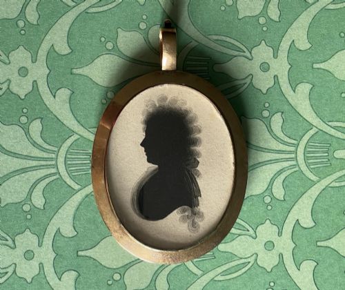 fig 1. A double-sided oval gold pendant containing two Georgian black painted silhouette profile portraits of a man and a woman set against off-white paper backgrounds.