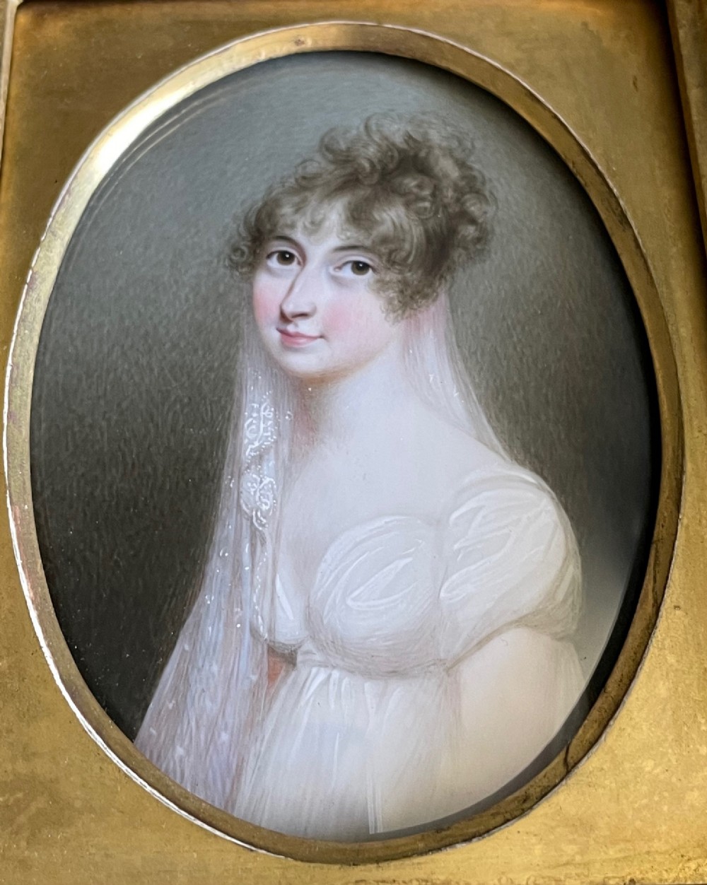 An oval watercolour miniature portrait depicting a woman with dark eyes, curled brown hair, and a translucent veil against a hatched grey background. View III