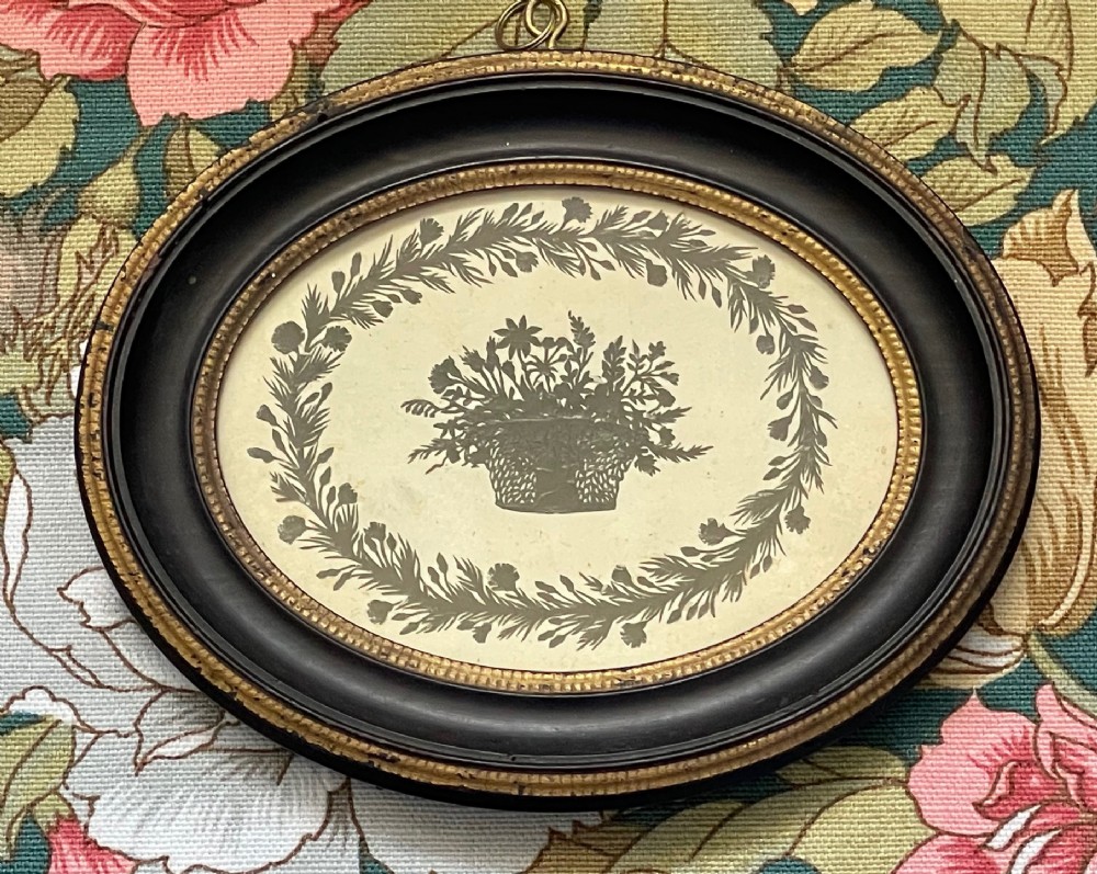 An oval framed Regency cut paper picture featuring a central basket of flowers enclosed by a leafy wreath set inside an ebonised frame with a gilt inner slip.
