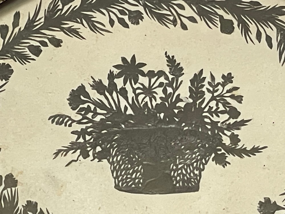 An oval framed Regency cut paper picture featuring a central basket of flowers enclosed by a leafy wreath set inside an ebonised frame with a gilt inner slip. View II