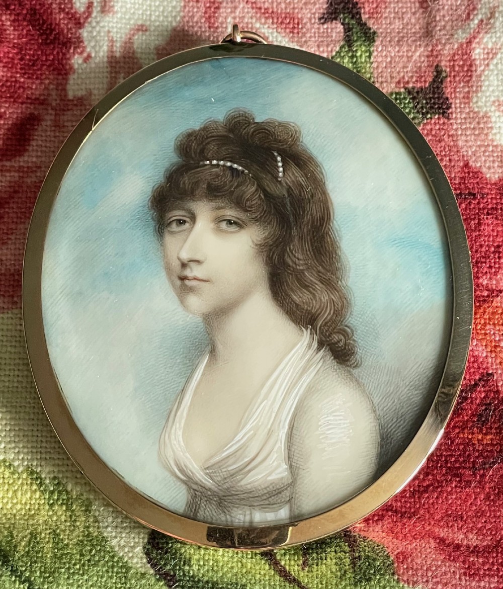An oval Georgian portrait miniature depicting a woman with brown curly hair adorned with pearls, wearing a white dress set against a sky blue background inside a gold frame.