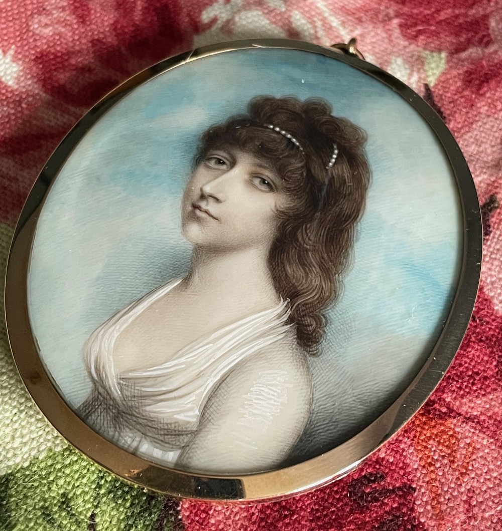 An oval Georgian portrait miniature depicting a woman with brown curly hair adorned with pearls, wearing a white dress set against a sky blue background inside a gold frame. View II