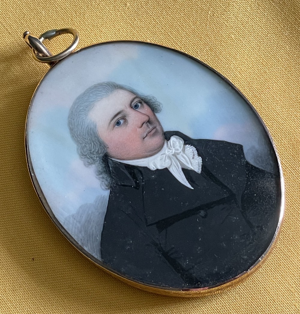 An oval Georgian portrait miniature depicting a gentleman in a black coat and white cravat set in a gold frame with a suspension ring.