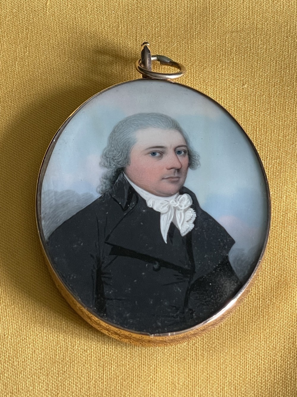 An oval Georgian portrait miniature depicting a gentleman in a black coat and white cravat set in a gold frame with a suspension ring. View II
