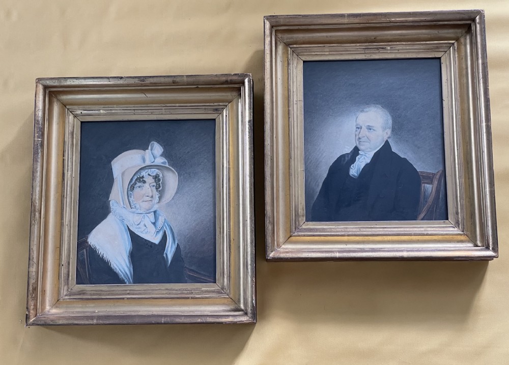 A pair of early 19th-century framed watercolour portraits depicting a man in a dark coat and a woman in a bonnet, housed in moulded giltwood frames.