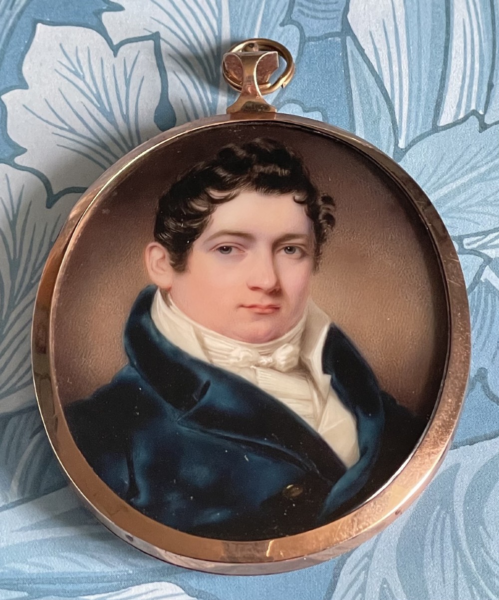 A circular antique portrait miniature of a gentleman in a dark blue coat and white cravat, encased in a gold-toned metal pendant frame.