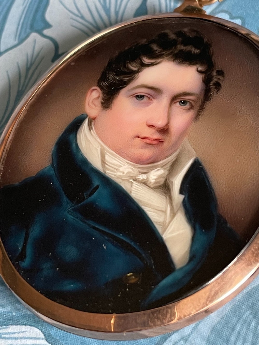 A circular antique portrait miniature of a gentleman in a dark blue coat and white cravat, encased in a gold-toned metal pendant frame. View II