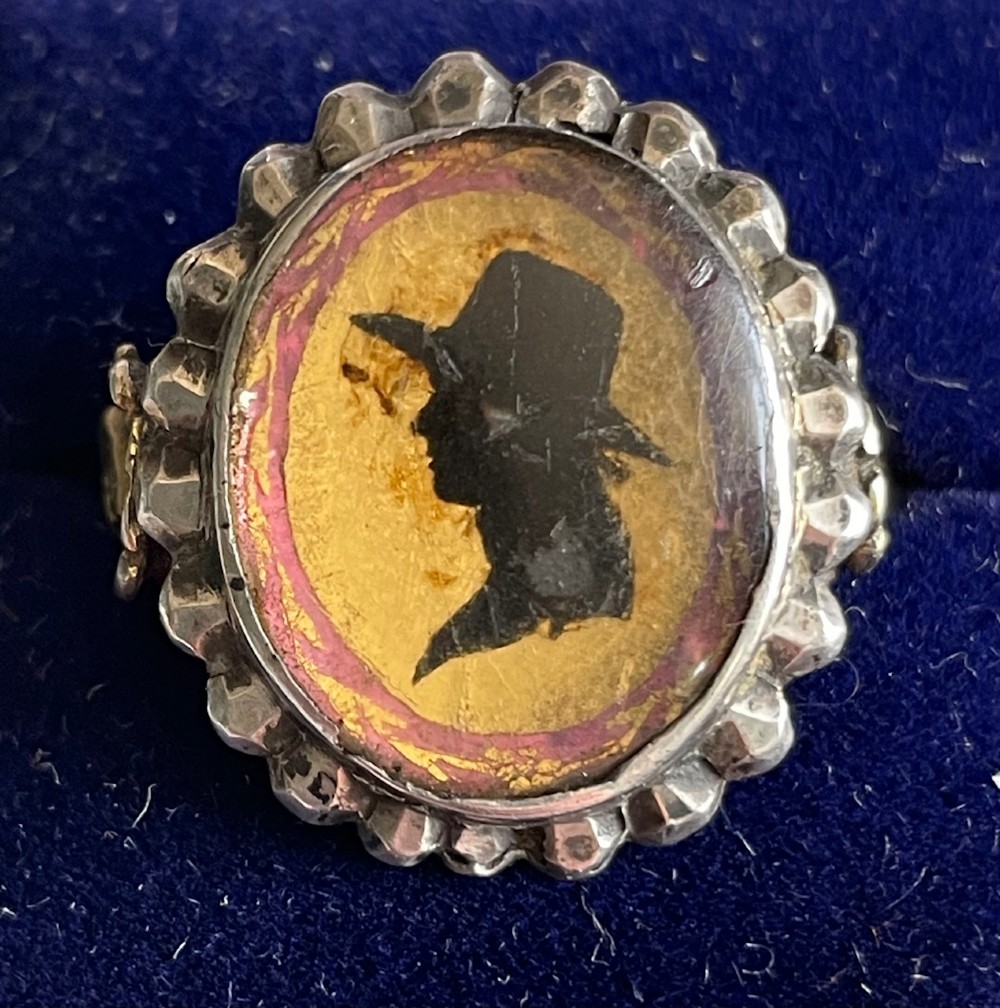 An oval antique Georgian silver ring featuring a black silhouette profile portrait of a figure wearing a hat set over a gold foil background within a crimped silver bezel.