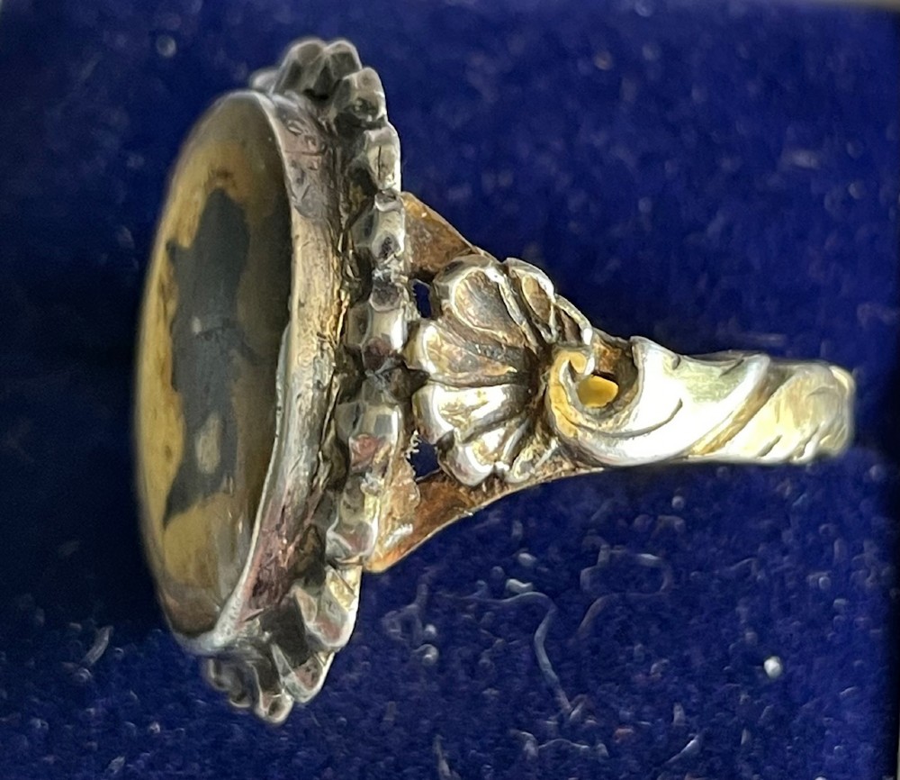 An oval antique Georgian silver ring featuring a black silhouette profile portrait of a figure wearing a hat set over a gold foil background within a crimped silver bezel. View II