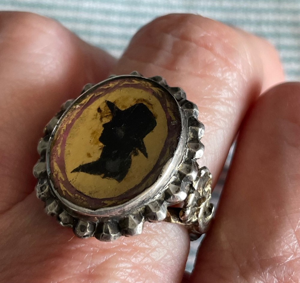 An oval antique Georgian silver ring featuring a black silhouette profile portrait of a figure wearing a hat set over a gold foil background within a crimped silver bezel. View IV