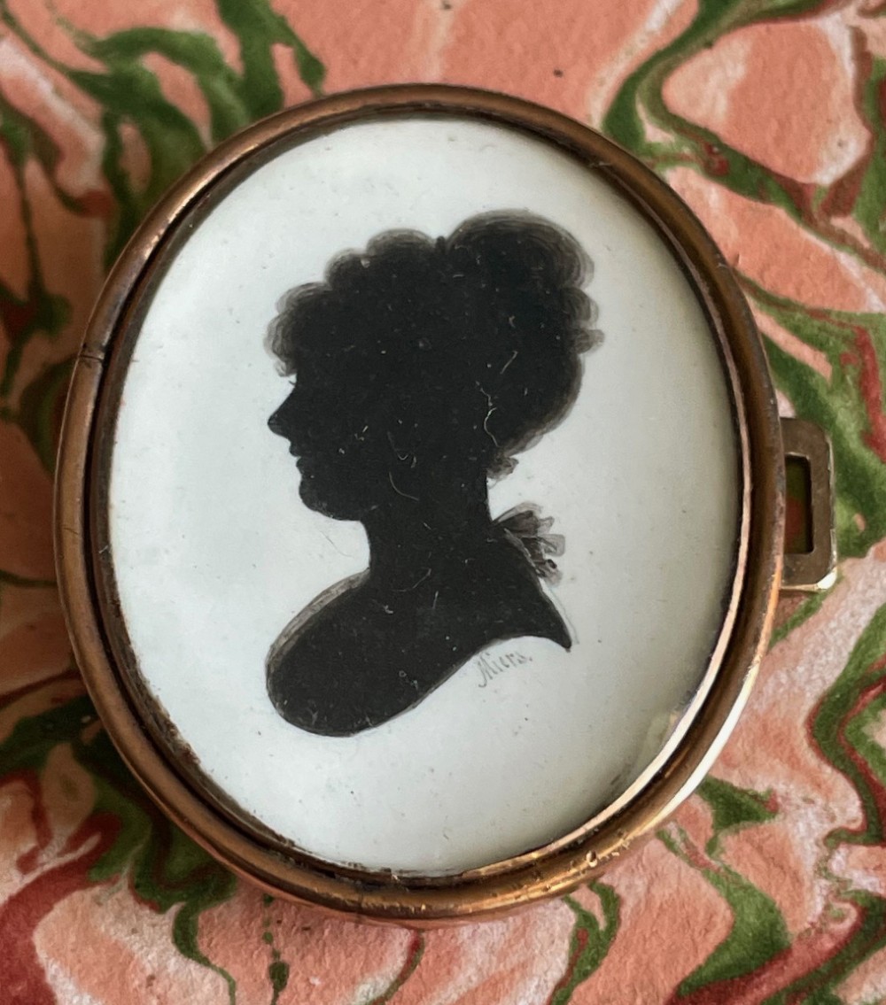 An oval Georgian gold-framed pendant featuring a black silhouette profile portrait of a woman against a light background, signed Miers.