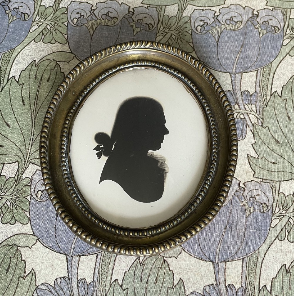 An oval brass framed profile silhouette portrait of a gentleman in a queue hairstyle set against a light background. View II