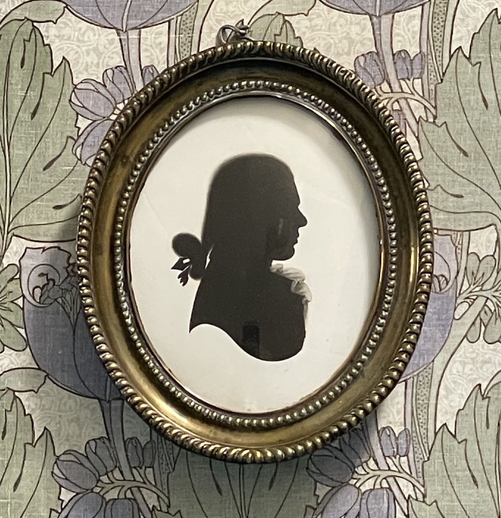 An oval brass framed profile silhouette portrait of a gentleman in a queue hairstyle set against a light background.