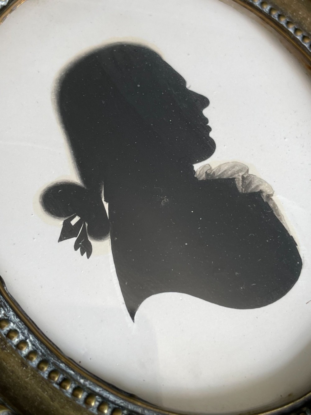 An oval brass framed profile silhouette portrait of a gentleman in a queue hairstyle set against a light background. View III