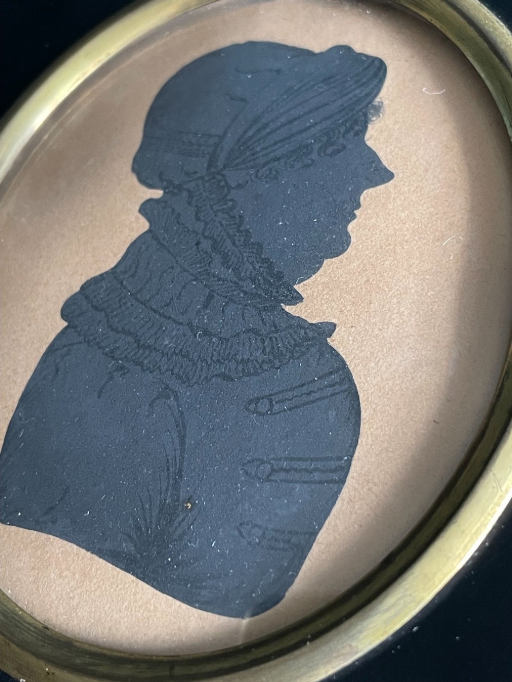 A profile silhouette portrait enclosed in an oval brass bezel and mounted within a rectangular black ebonised frame featuring a brass acorn hanger. View II