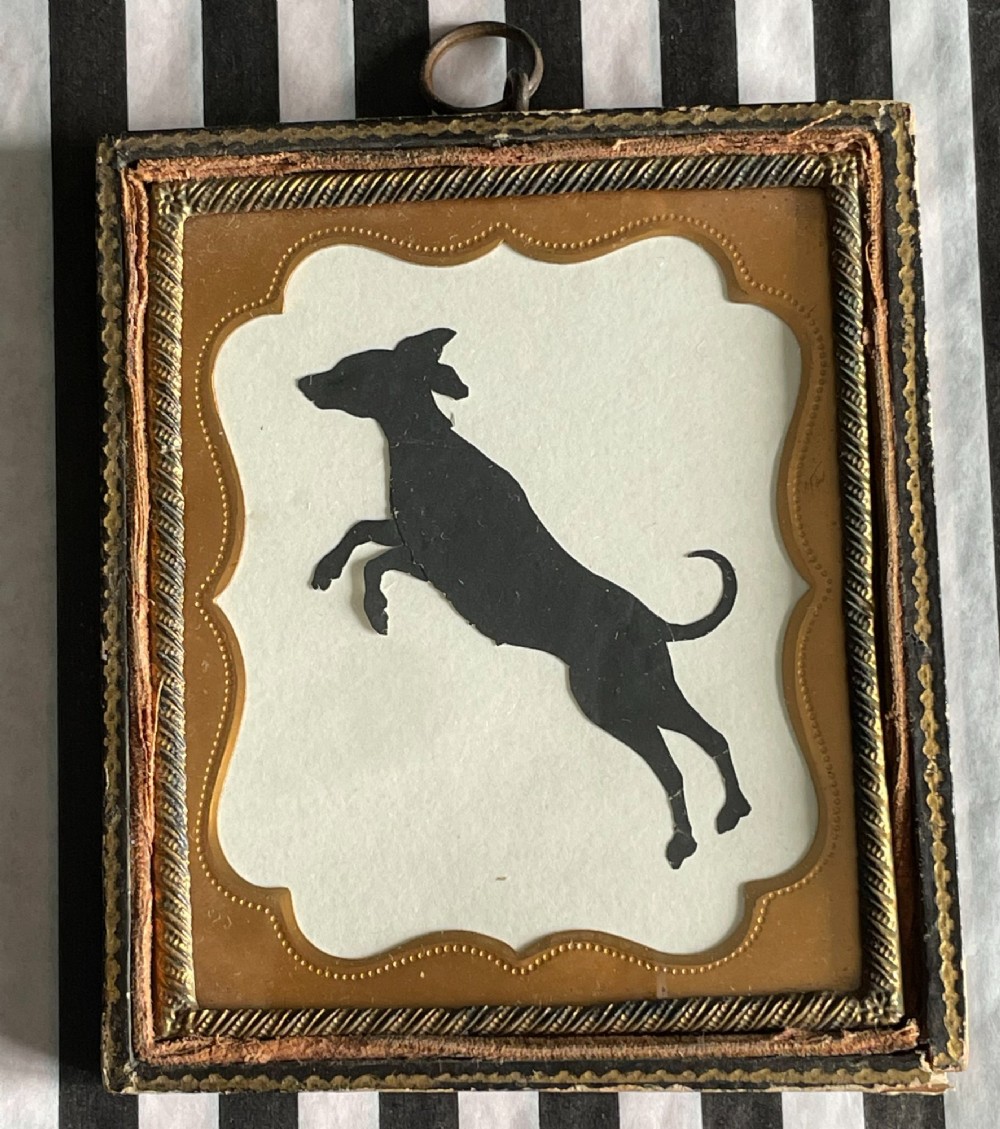 A framed nineteenth-century black cut-paper silhouette depicting a leaping dog, mounted inside an embossed gold-toned brass frame with a top suspension ring.