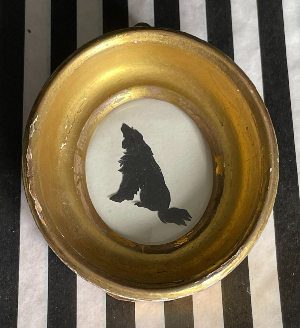 A miniature black cut-paper silhouette depicting a seated dog profile within a deep, oval giltwood frame.