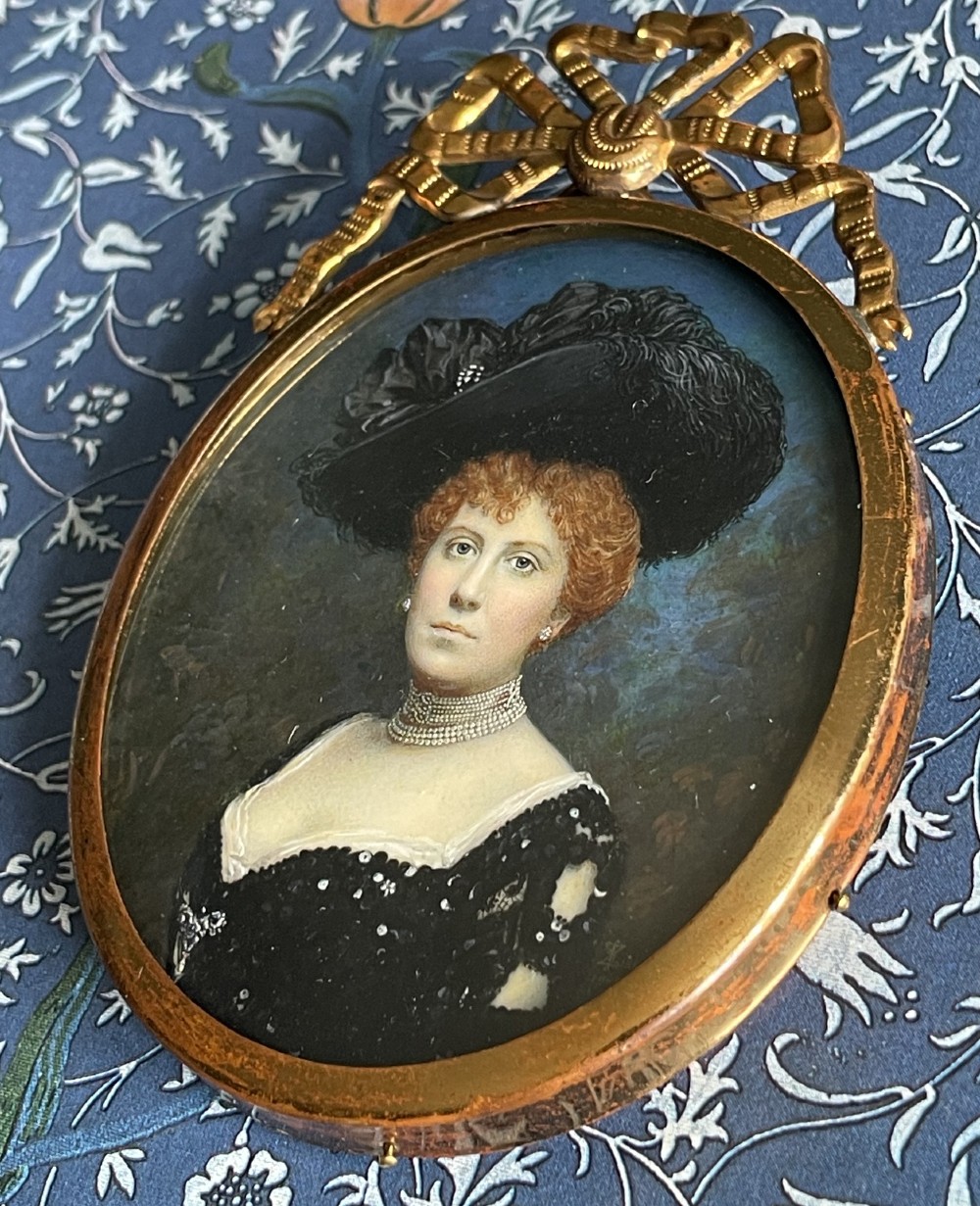 An oval Edwardian portrait miniature depicting a woman with reddish hair in a black feather hat, encased in a gilt brass frame with a ribbon bow crest. View V