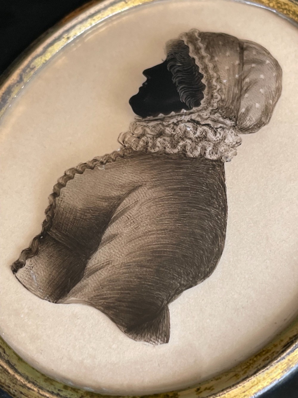 A profile silhouette portrait of a woman in a bonnet painted on glass, mounted in an oval brass bezel within a rectangular black papier-mache frame with an acorn hanger. View II