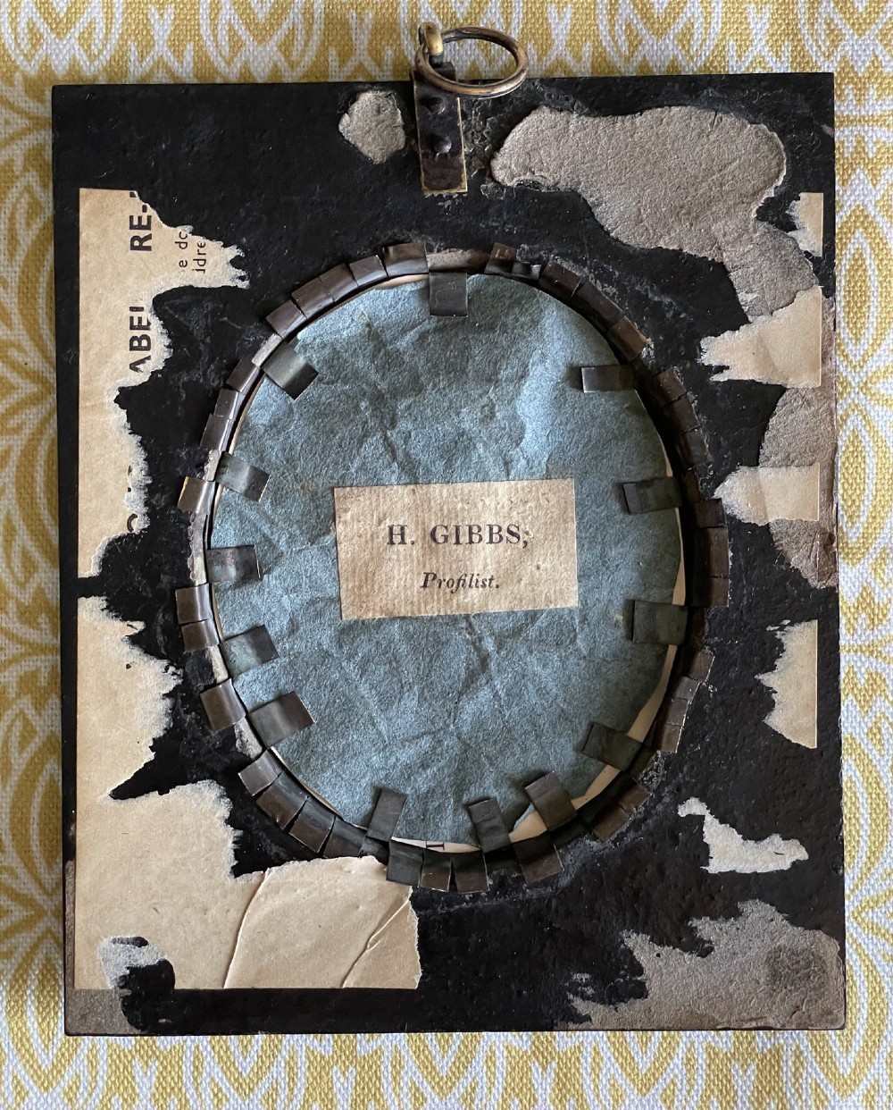 A profile silhouette portrait of a woman in a bonnet painted on glass, mounted in an oval brass bezel within a rectangular black papier-mache frame with an acorn hanger. View III