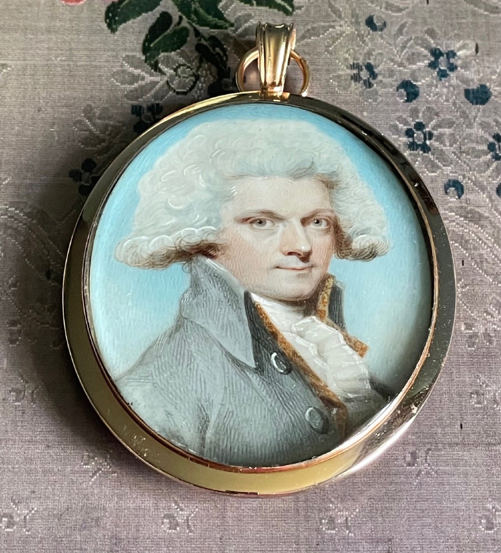 An oval Georgian portrait miniature depicting a gentleman with a white powdered wig and grey coat, set within a gold frame with a top loop.