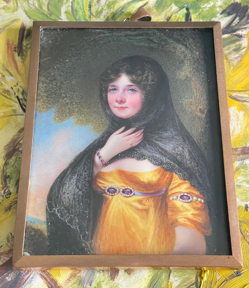 A Georgian enamel portrait miniature depicting a woman in a yellow gown and black lace head covering, presented in a metal frame with a top loop.