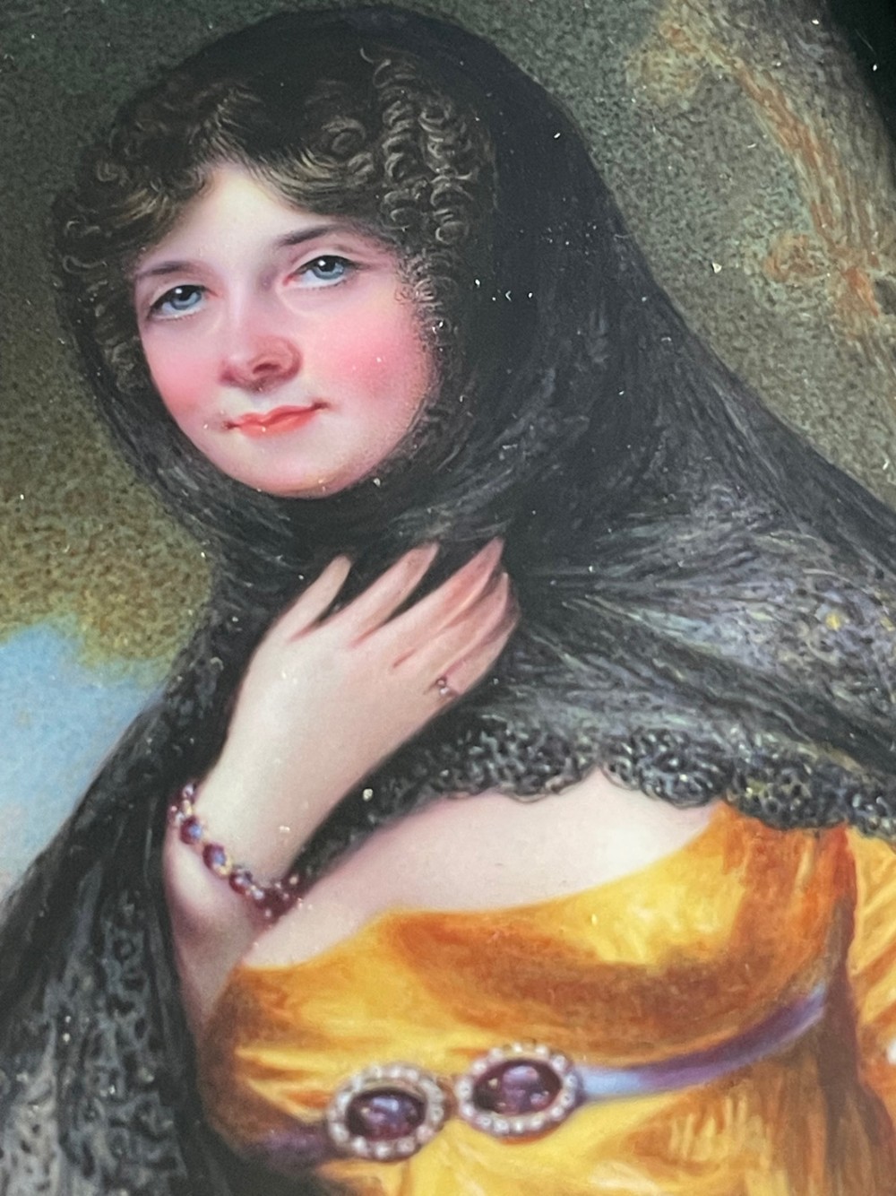 A Georgian enamel portrait miniature depicting a woman in a yellow gown and black lace head covering, presented in a metal frame with a top loop. View V