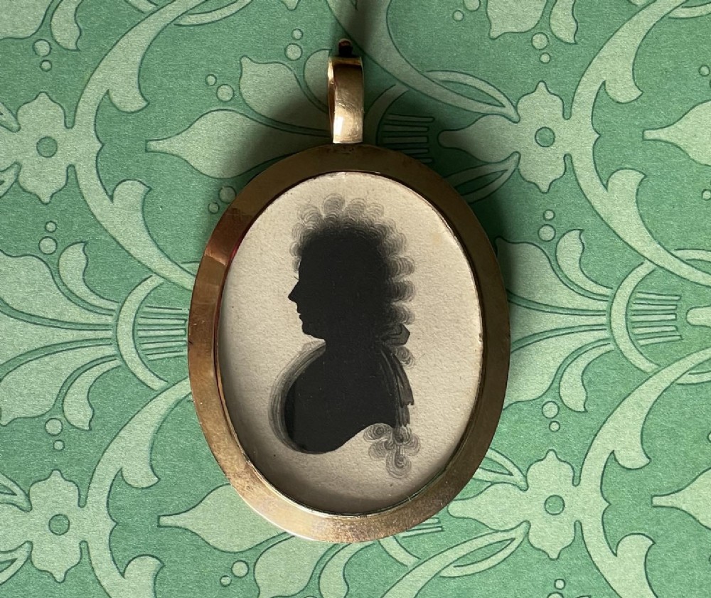 A double-sided oval gold pendant containing two Georgian black painted silhouette profile portraits of a man and a woman set against off-white paper backgrounds. View II