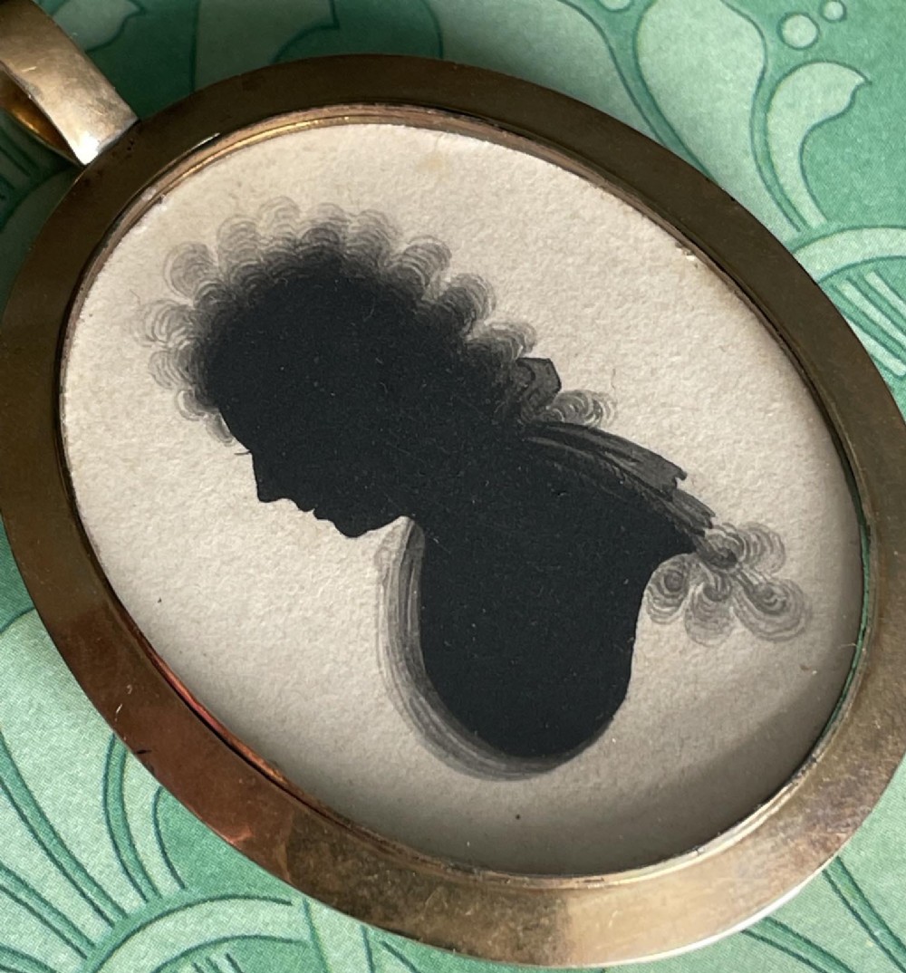 A double-sided oval gold pendant containing two Georgian black painted silhouette profile portraits of a man and a woman set against off-white paper backgrounds. View III