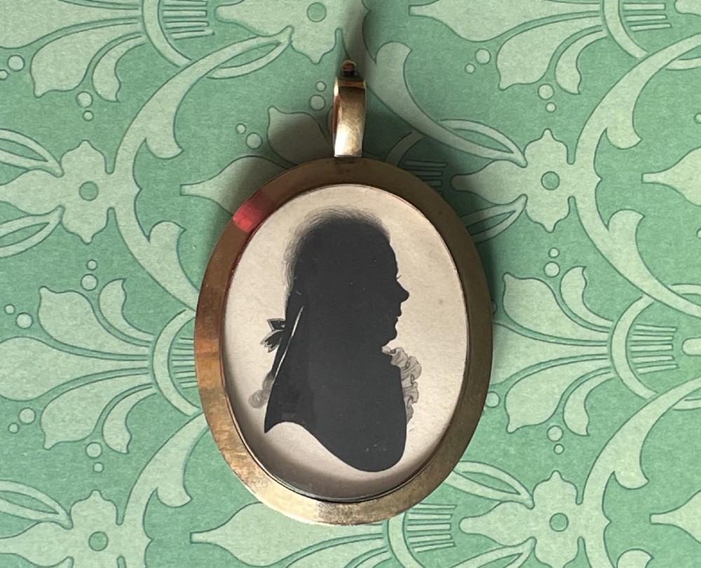 A double-sided oval gold pendant containing two Georgian black painted silhouette profile portraits of a man and a woman set against off-white paper backgrounds. View IV