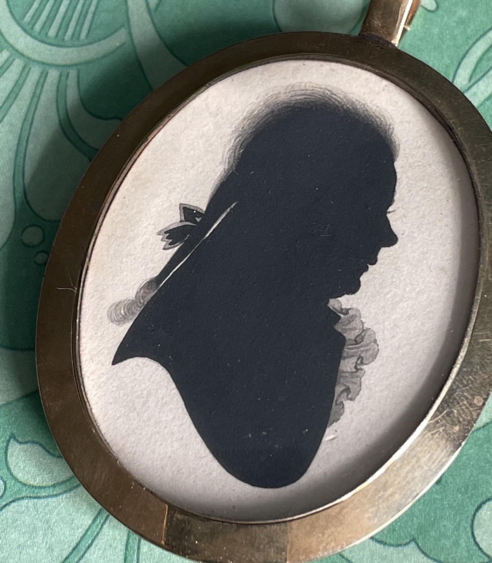 A double-sided oval gold pendant containing two Georgian black painted silhouette profile portraits of a man and a woman set against off-white paper backgrounds. View V