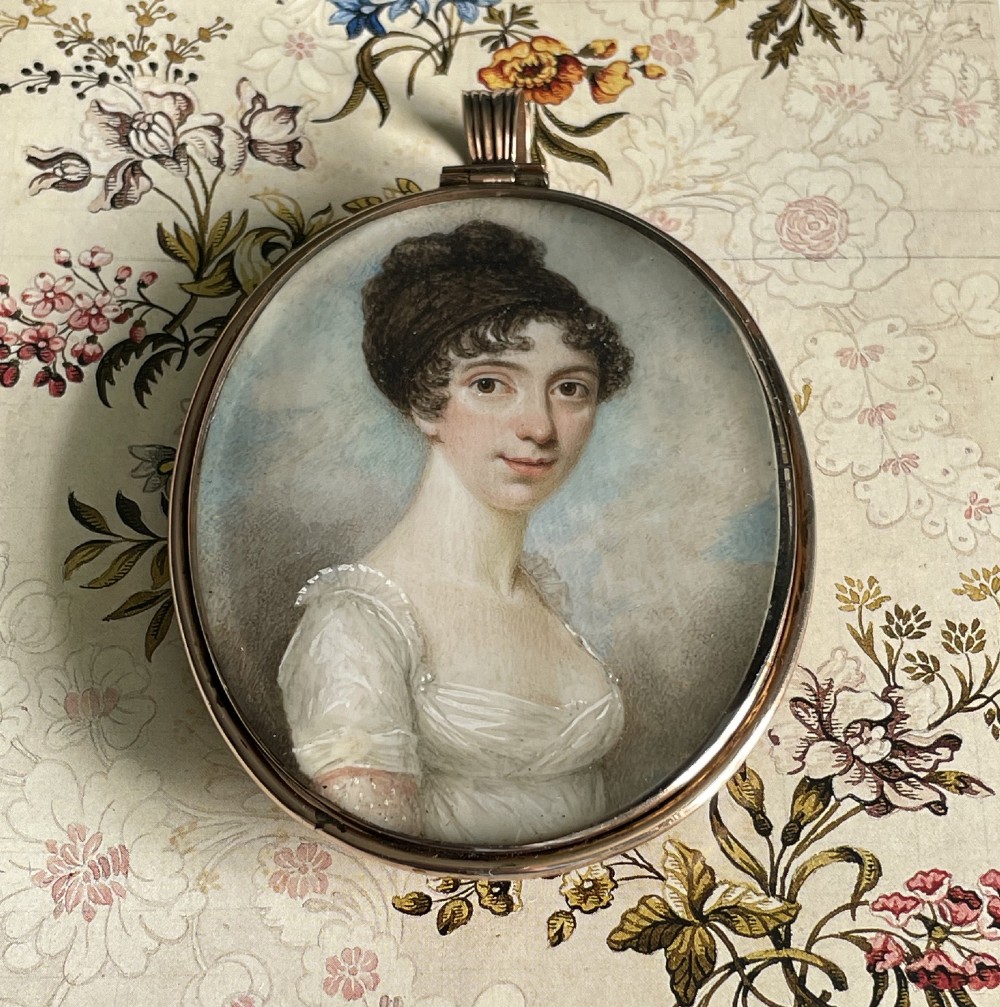 An oval Regency portrait miniature depicting a woman with dark hair in an updo wearing a white high-waisted gown, mounted in a gold casing with a ribbed loop.