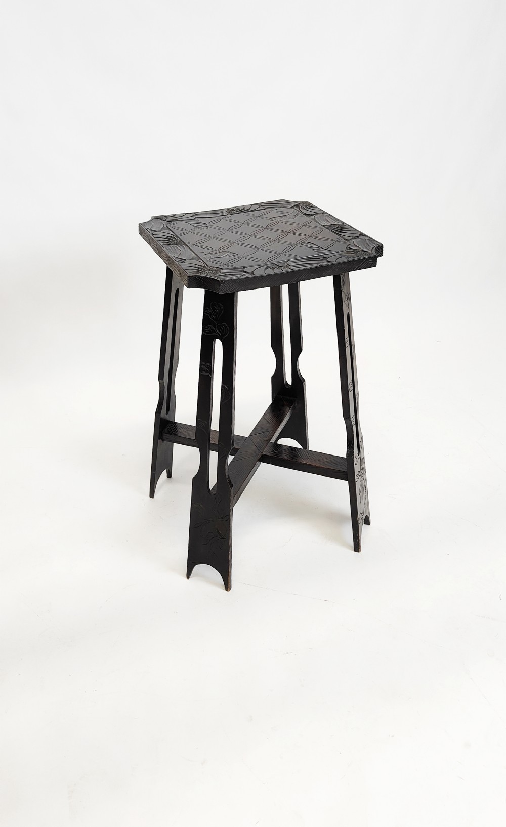 A square ebonised wooden side table featuring carved geometric and floral patterns on top and tapered legs joined by an X-frame stretcher.