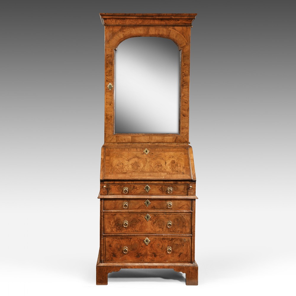 An antique William and Mary burr walnut bureau bookcase featuring an arched mirror upper door above a fall-front desk and chest of drawers with brass handles.
