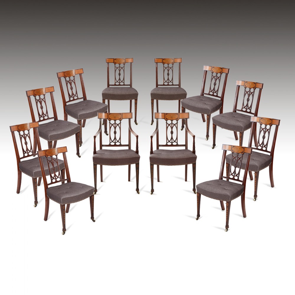 A set of twelve mahogany dining chairs featuring inlaid crest rails, pierced openwork splats, dark upholstered seats, and tapered legs with brass castors.