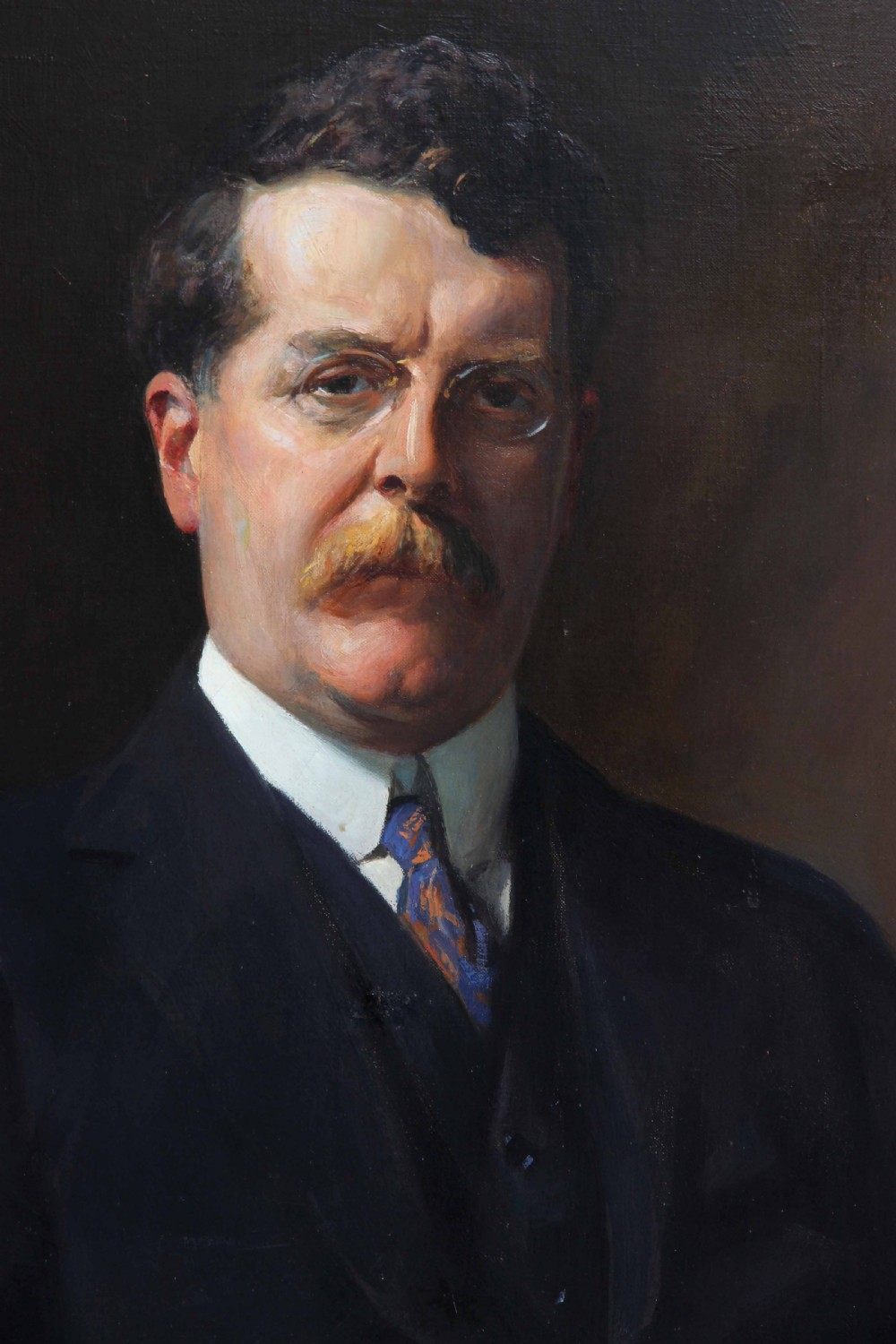 An antique oil painting depicting a seated gentleman wearing a dark suit and spectacles, set within an ornate gilded frame. View III